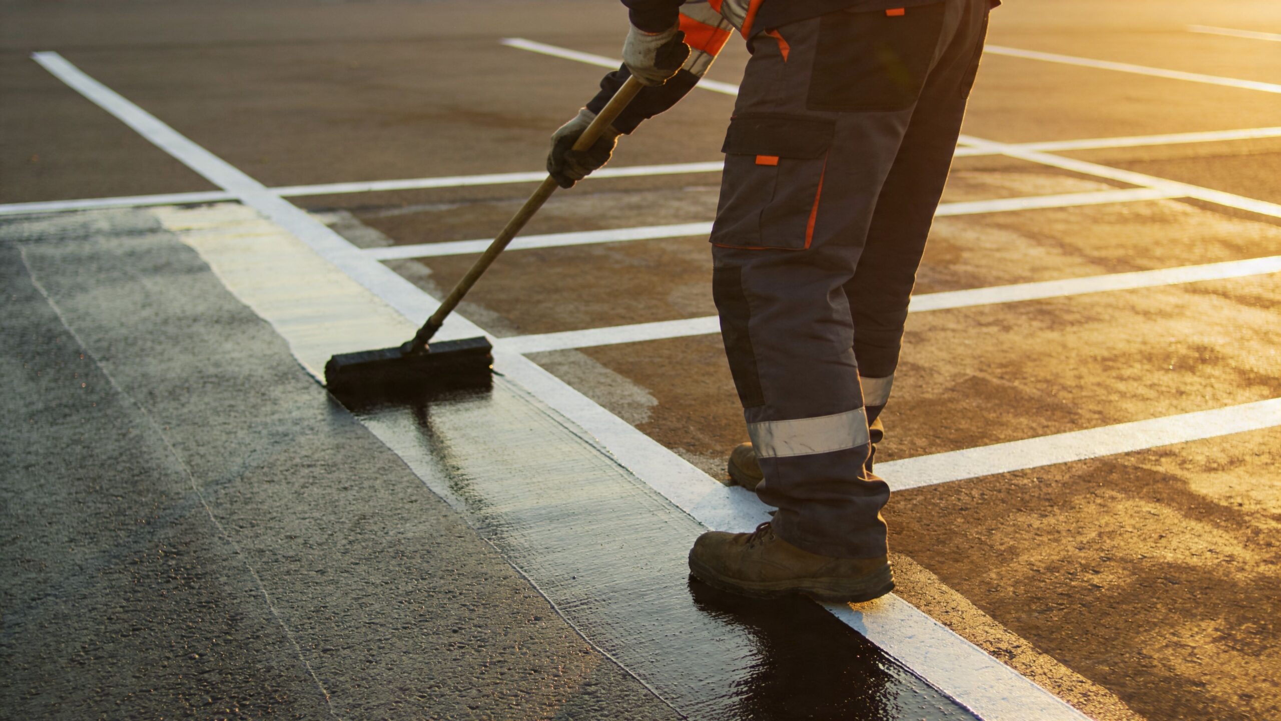 A worker applying dark sealant to a concrete slab at a parking lot during golden hour sunset.