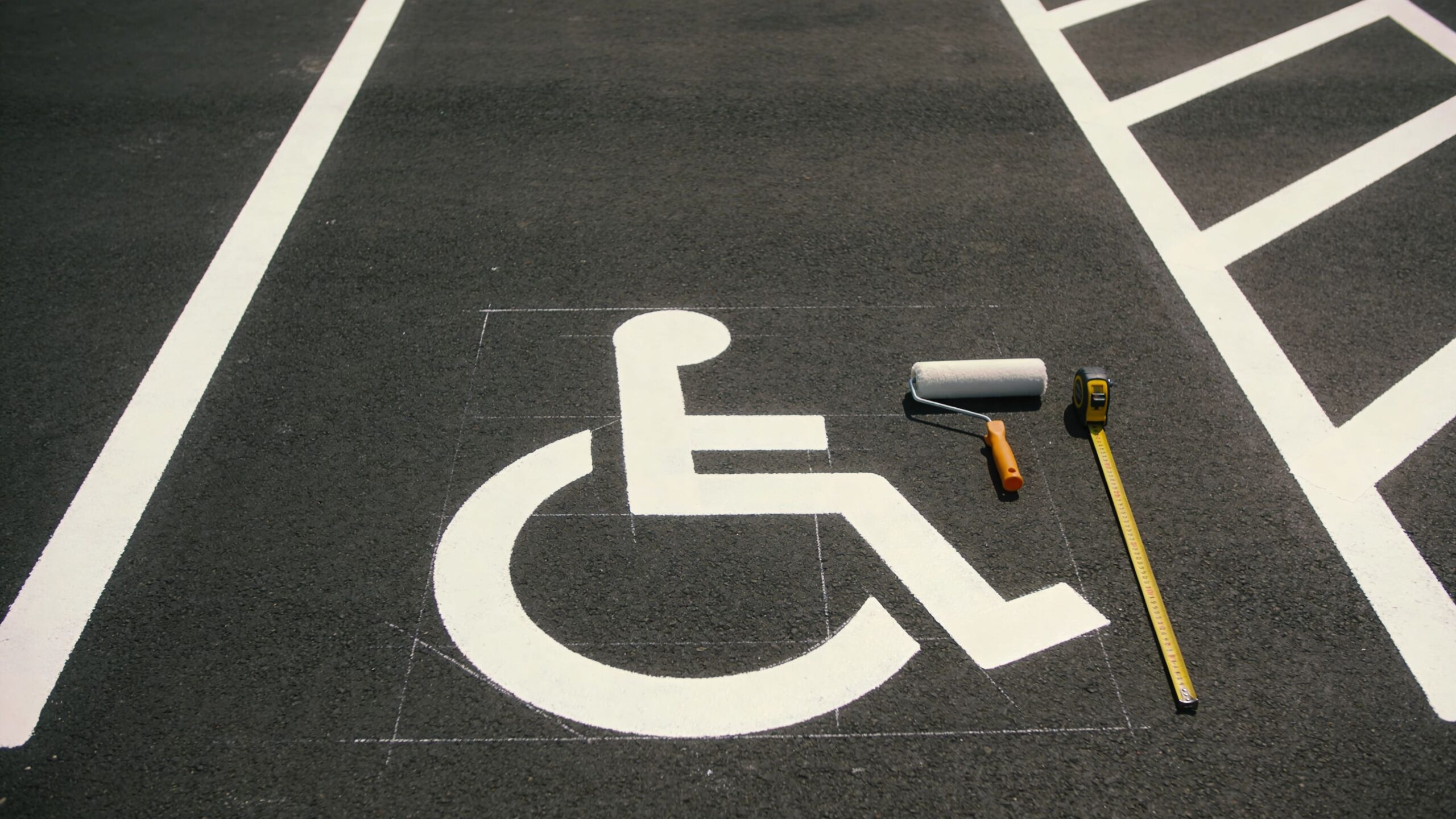 A fresh white handicap parking symbol painted on asphalt next to a paint roller and tape measure.
