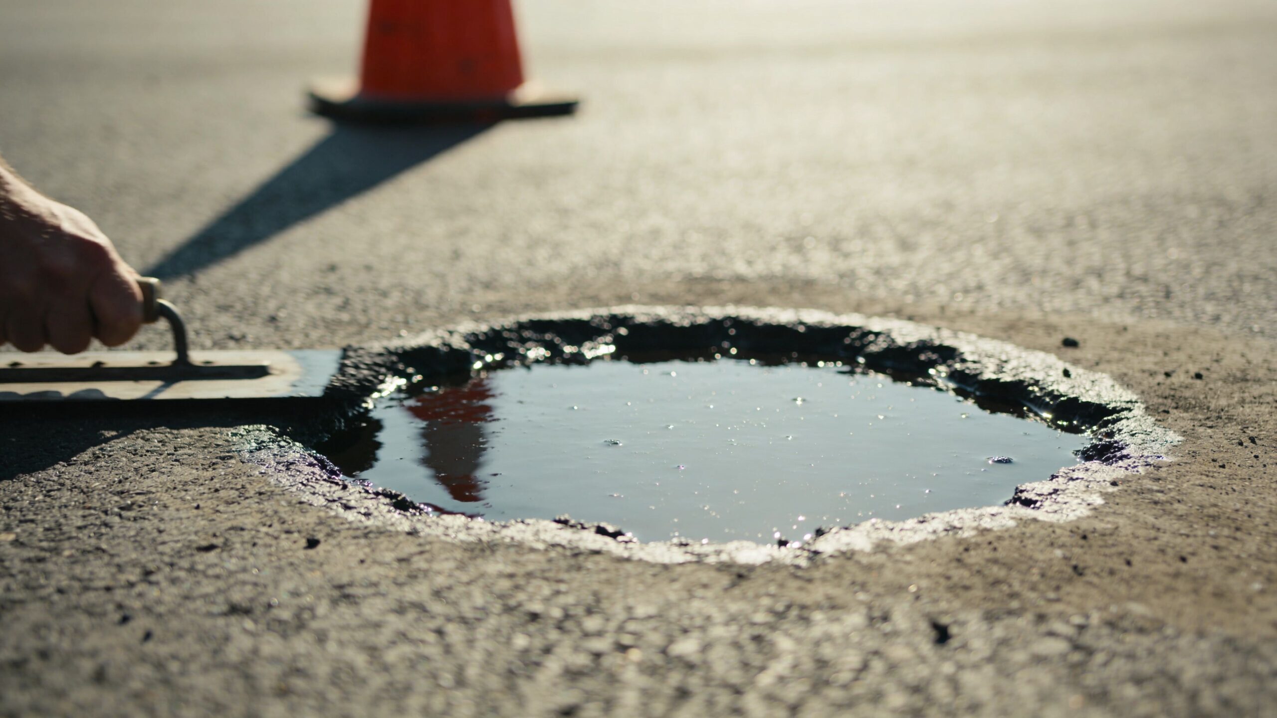 A construction worker uses a metal trowel to smooth out asphalt while repairing a pothole in a lot.